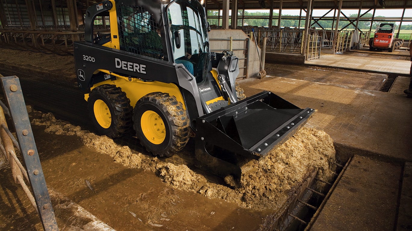 320D Skid Steer pushes material with a scraper attachment in a livestock shelter