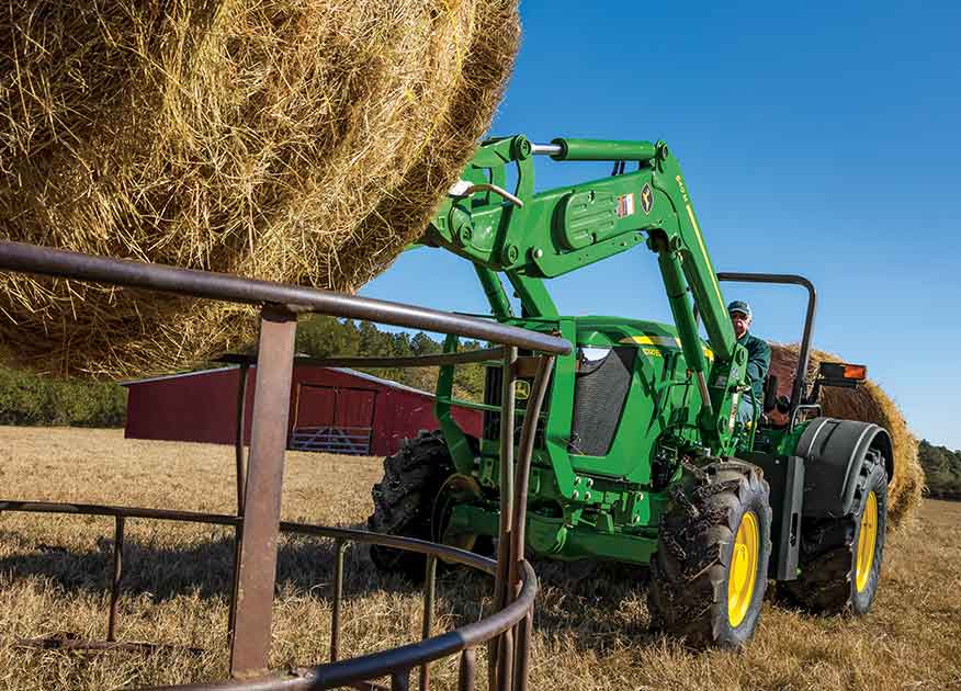 Side view of a John Deere Compact Tractor with fork attachments moving a hay barrel