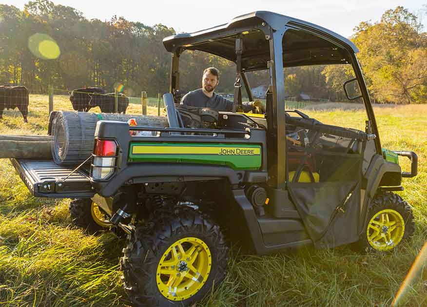 Man standing next to a John Deere Gator Utility Vehicle with equipment in the bed and cattle in the background