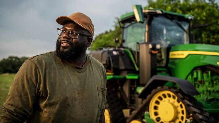 A black farmer standing proudly near a John Deere tractor