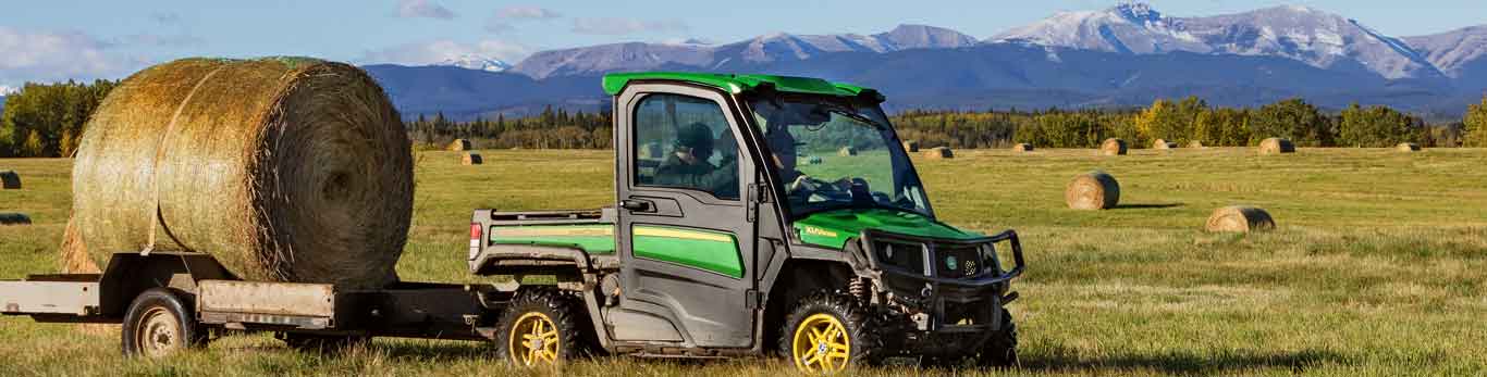 Two people driving a John Deere Gator Utility Vehicle through a field with a hay bale on a trailer