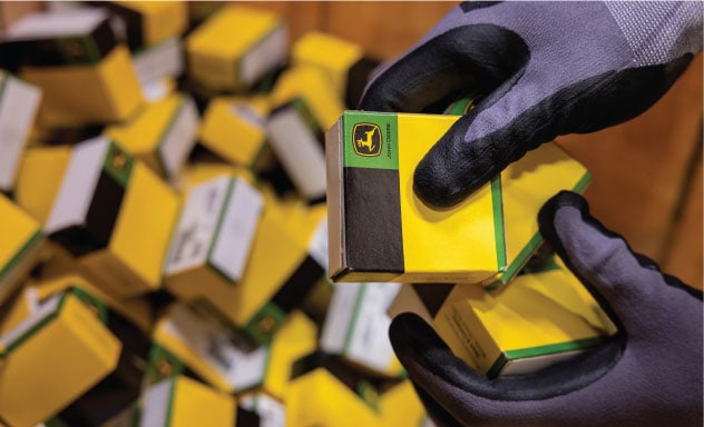John Deere small cardboard box is held by a person with gloves above a box full of smaller parts boxes.