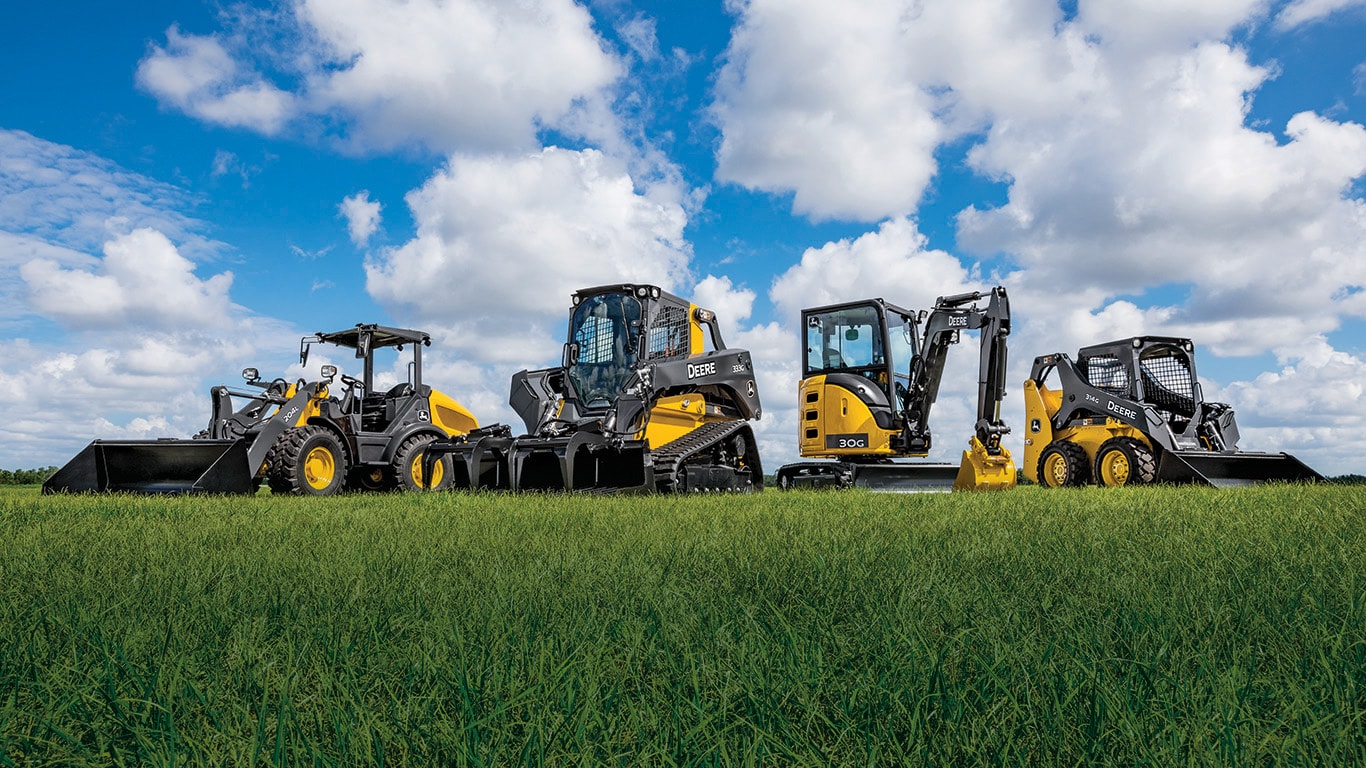 Multiple pieces of John Deere compact construction equipment parked in grassy field.