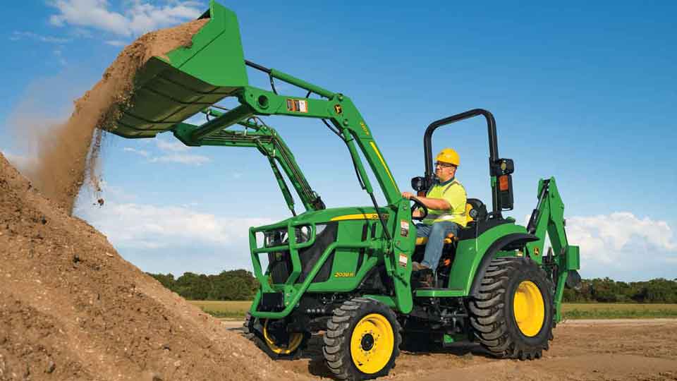 Man with yellow vest and hard hat operating a John Deere 2038R Compact Tractor and dumping dirt out of the bucket