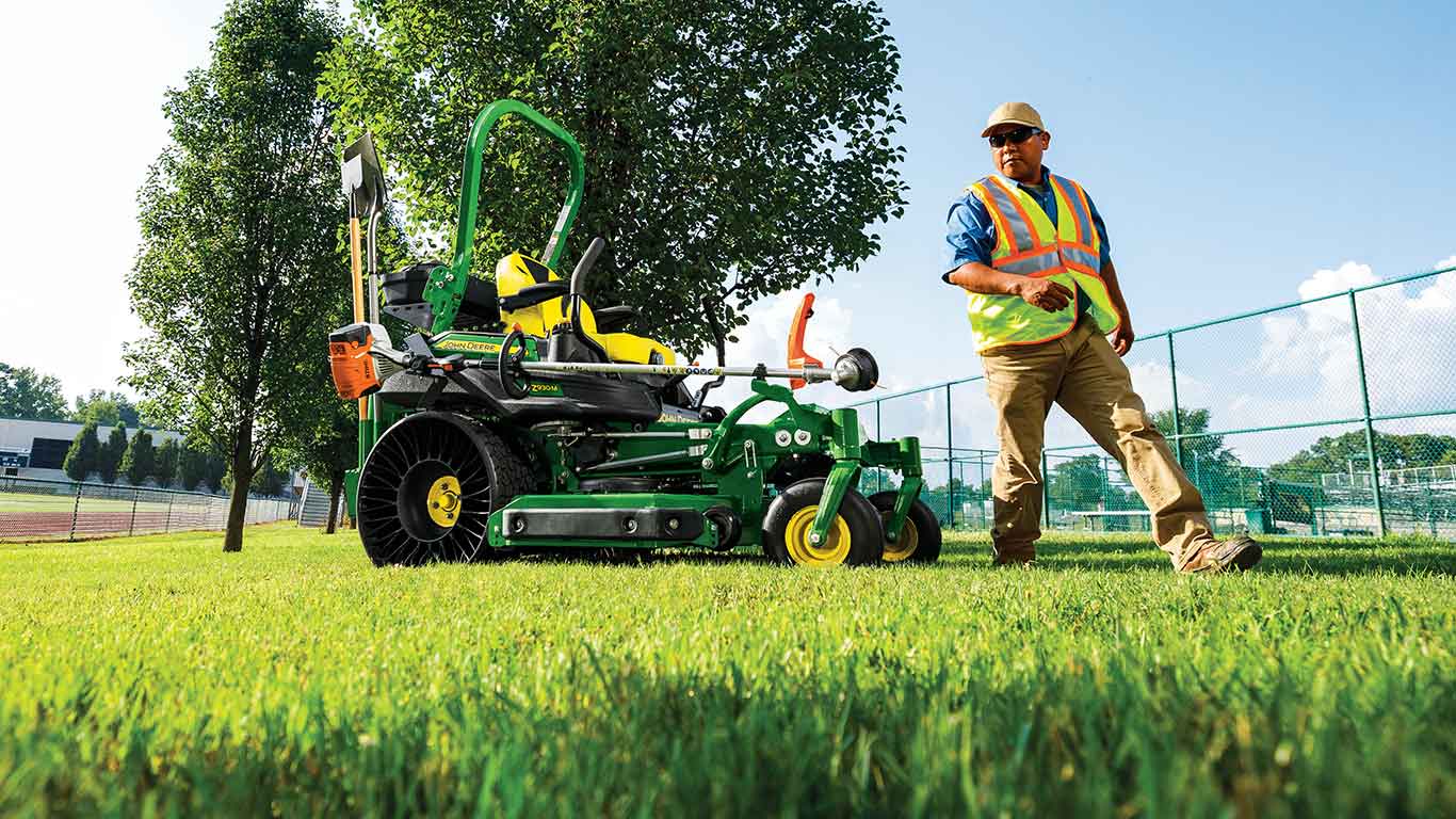 John Deere Zero Turn mower with trimmer and shovels attached with man in yellow vest walking by