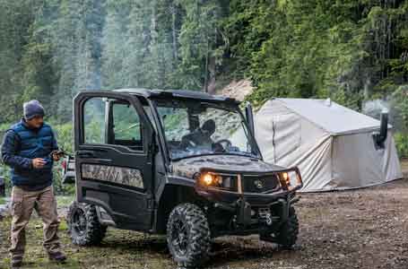 Camouflage John Deere Gator Utility Vehicle in a wooded area with one man inside and another standing outside and a tent in the background 