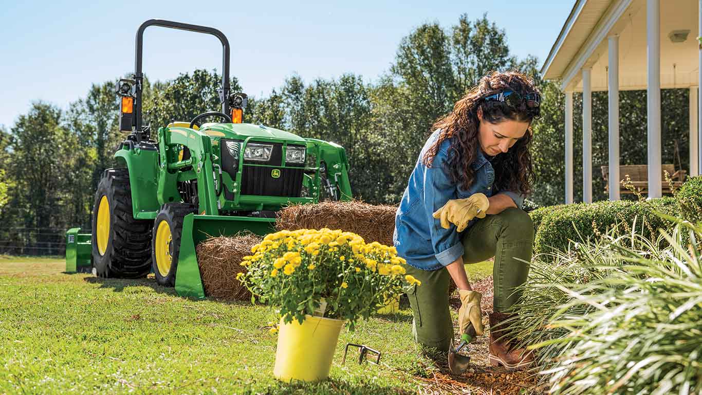 Woman gardening in a yard with a John Deere Compact Tractor with bucket attachment sits in the background