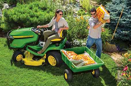 Woman sitting on a John Deere Lawn Tractor in a yard with a man loading fertilizer to the attached poly cart