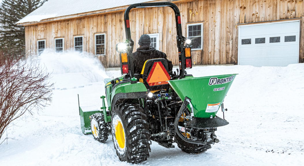Person clearing snow on a 2038R compact tractor with snowblower and Frontier spreader.