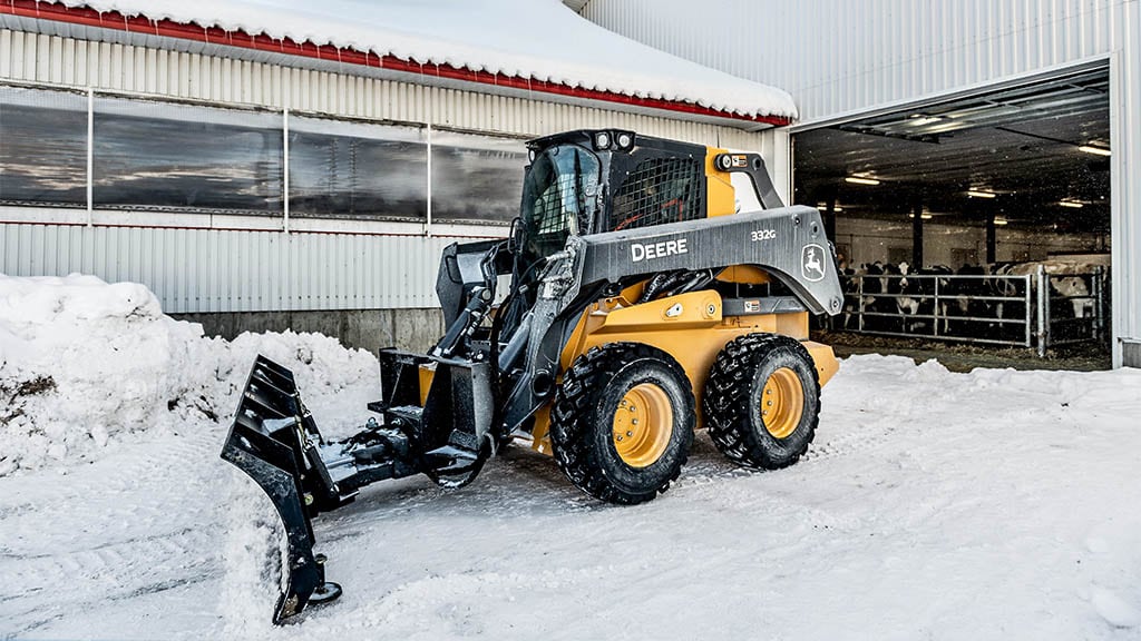 Person using skid steer equipped with a blade to clear snow in front of barn