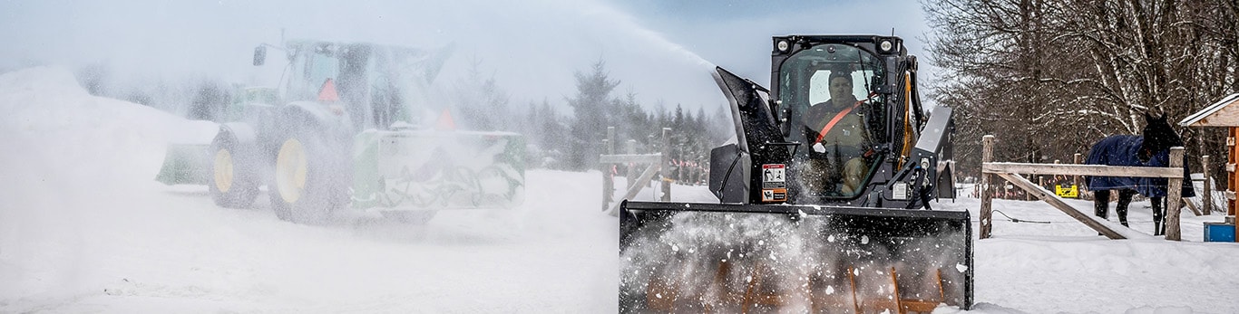 Person blowing snow using a skidsteer and snowblower with a 5 series utility tractor equipped with a snow blower and loader in the background.